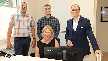 Landrat Jan Weckler, Alexander Hiemer, Schulleiterin Claudia Baumann und Jan Klüners in einem modernen Raum. Sie stehen um einen Schreibtisch herum.