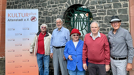 Das Bild zeigt fünf Personen vor einger Steinmauer neben einem Roll-Up des Vereins KULTURplus.