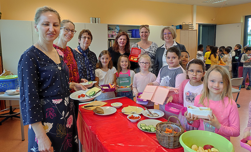 Kinder und Erwachsene stehen um einen Tisch. Darauf sind mehrere Teller mit Obst, Gemüse, Käse und Brot.