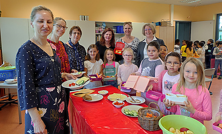 Kinder und Erwachsene stehen um einen Tisch. Darauf sind mehrere Teller mit Obst, Gemüse, Käse und Brot.