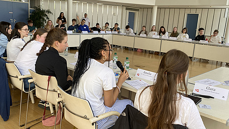 Junge Leute an Tischen. Eine Jugendliche spricht in ein Mikrofon.