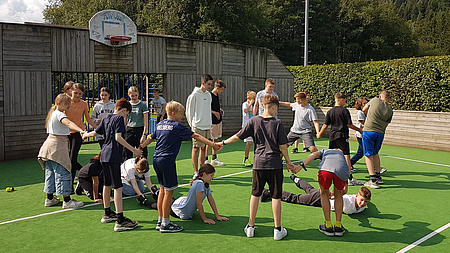 Eine Gruppe Jugendlicher steht im Kreis auf einem Sportfeld. Sie spielen ein Spiel.