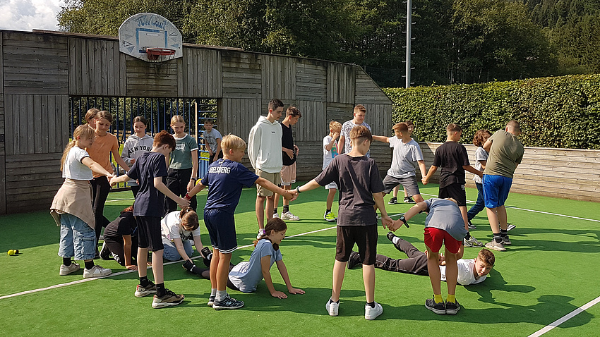 Eine Gruppe Jugendlicher steht im Kreis auf einem Sportfeld. Sie spielen ein Spiel.