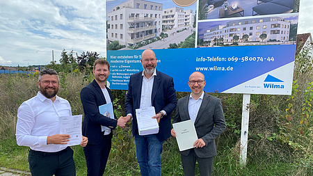 Vier Männer stehen nebeneinander. Jeder von ihnen hat ein Schriftstück in der Hand, die beiden Männer in der Mitte geben sich die Hand. Im Hintergrund ein Bauschild und eine Wiese.