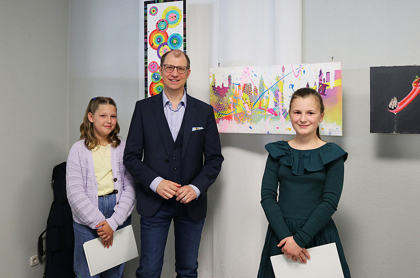 Ein Mann und zwei Mädchen, die Urkunden in der Hand halten. Im Hintergrund hängen Bilder an der Wand.