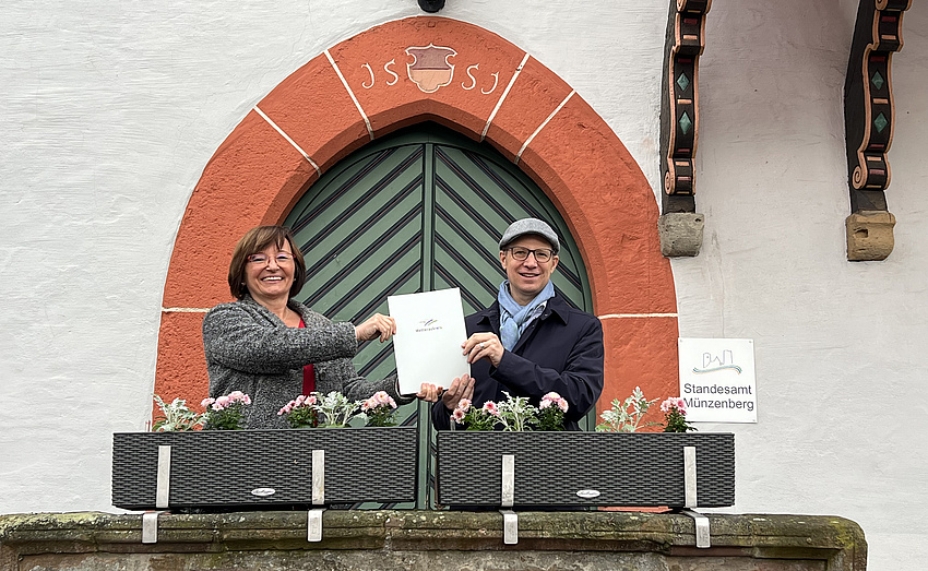 Ein Mann und eine Frau halten gemeinsam ein Schriftstück in Händen. Sie stehen auf einer Art Balkon, davor zwei Blumenkästen, im Hintergrund eine Holztür im gotischen Baustil mit Spitzbogen.
