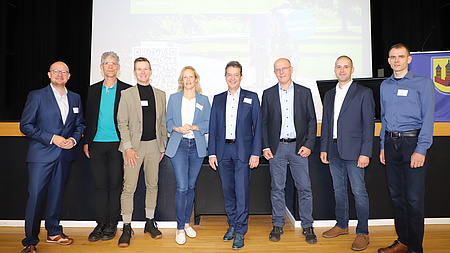 Eine Frau und sieben Männer stehen in seinem Saal vor einer Bühne.