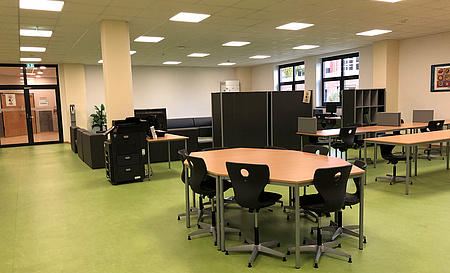 Ein großer Raum mit verschiedenen Sitzgelegenheiten. Runde Tische mit Stühlen und Sofaecken. Dazwischen Regale.