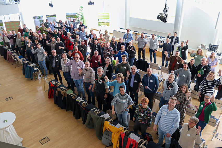 Eine große Gruppe von etwa 80 Personen steht in einem Raum. Alle lächeln und winken in die Kamera.