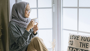 Frau mit Kopftuch sitzt mit einer Tasse an einem Fenster