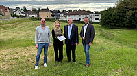 Eine Frau und drei Männer stehen gemeinsam auf einer Wiese an einem Ortsrand. Die Frau und einer der Männer halten gemeinsam ein Dokument in Händen.