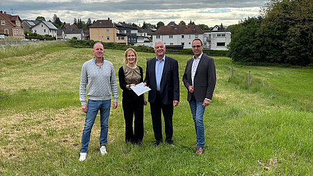 Eine Frau und drei Männer stehen gemeinsam auf einer Wiese an einem Ortsrand. Die Frau und einer der Männer halten gemeinsam ein Dokument in Händen.