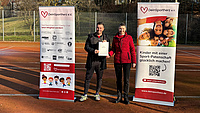 Eine Frau und ein Mann stehen auf einem Tennisplatz im Freien und halten gemeinsam ein Dokument in Händen. Links und rechts von ihnen stehen zwei Plakate.