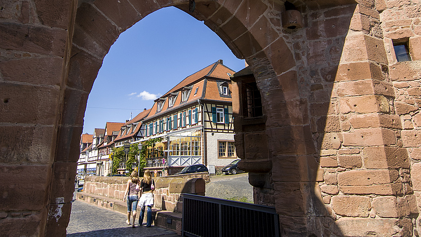 Zwei Frauen laufen durch einen mittelalterlichen Torbogen über eine Brücke. Im Hintergrund sieht man ein Fachwerkhaus.