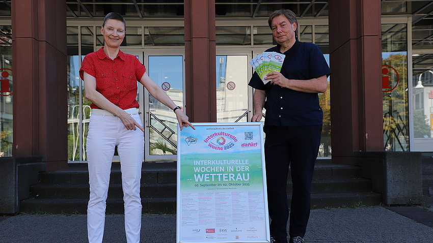 Eine Frau und ein Mann stehen vor einem Gebäude. Zwischen beiden befindet sich ein Plakat, auf das die Frau zeigt. Der Mann hält mehrere Flyer in der Hand.
