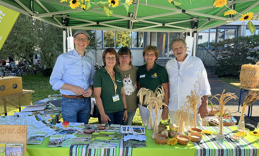 Drei Frauen und zwei Männer an einem Marktstand unter einem Sonnenschirm.
