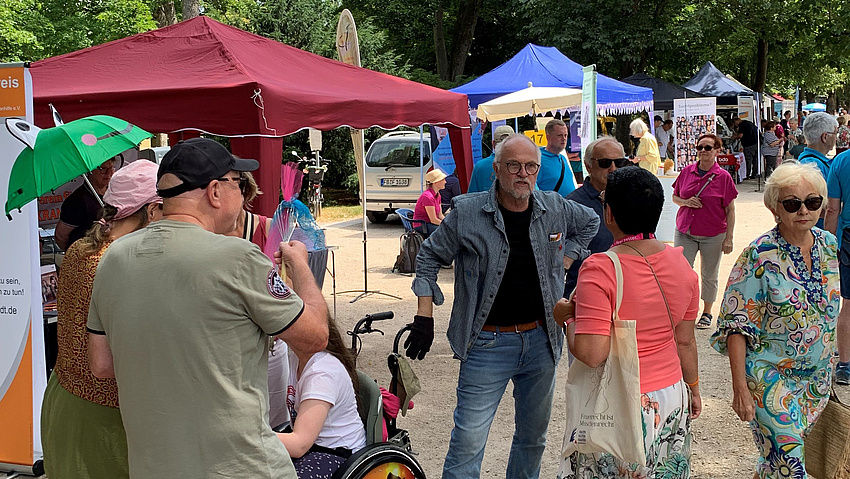 Auf einem Parkgelände sind mehrere Stände zu sehen und Personen, die sich an ihnen informieren oder miteinander sprechen.