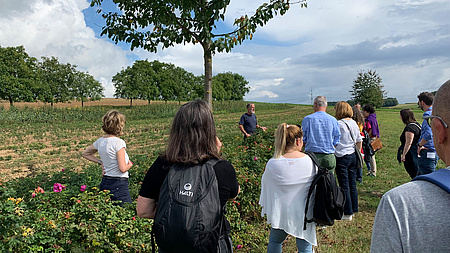 Personengruppe steht auf einem Feld. Im Vordergrund Blumen, ein Obstbaum, im Hintergrund am Feldrand stehen Bäume.