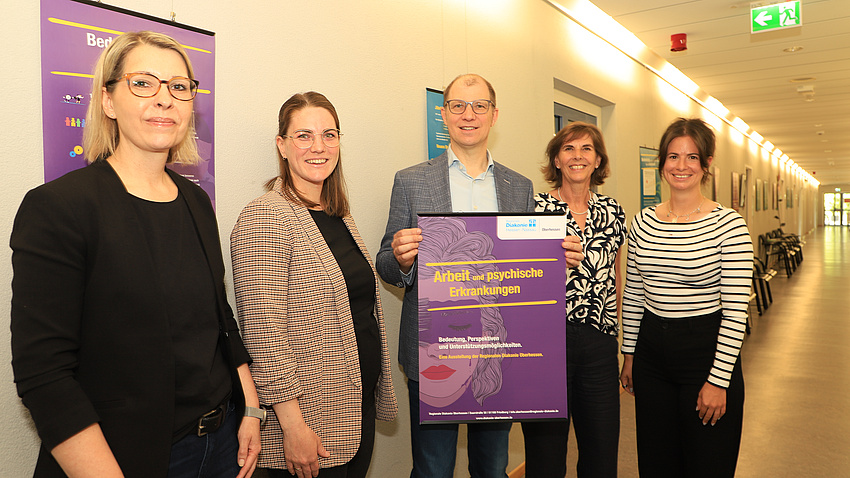 Vier Frauen und ein Mann stehen in einem langen Flur, an dessen Wänden mehrere Plakate hängen. Der Mann hält zudem ein weiteres Plakat in die Höhe.