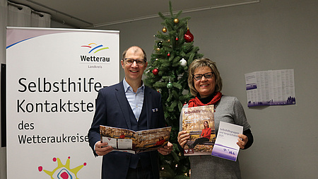 Zwei Menschen stehen mit jeweils einer Zeitschrift in der Hand vor einem Tannenbaum.