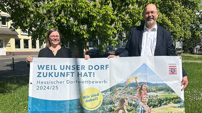 Ein Mann und eine Frau stehen auf einer Wiese. Sie halten ein Banner in die Kamera. Darauf steht: "Weil unser Dorf Zukunft hat!"