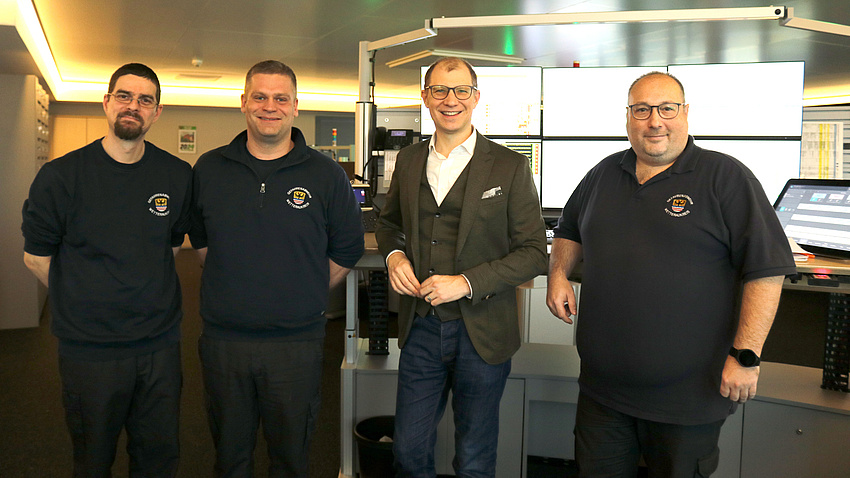 Vier Männer stehen nebeneinander. Im Hintergrund Computerbildschirme.