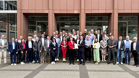 Zahlreiche Personen stehen vor dem Eingangsbereich eines Gebäudes. Zwei von ihnen, eine Frau und ein Mann in der Mitte dieser Gruppe, halten ein Dokument in die Höhe.