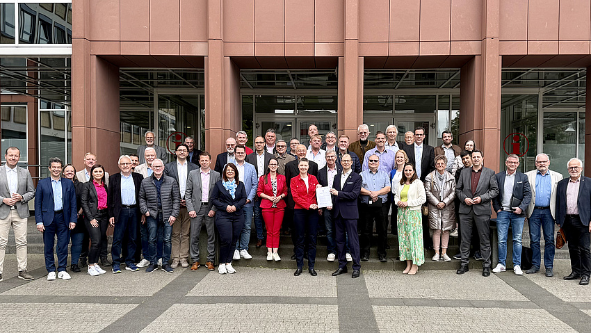 Zahlreiche Personen stehen vor dem Eingangsbereich eines Gebäudes. Zwei von ihnen, eine Frau und ein Mann in der Mitte dieser Gruppe, halten ein Dokument in die Höhe.