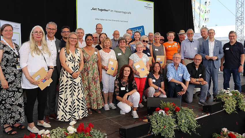 Zahlreiche Personen, Männer und Frauen, stehen auf einer Bühne im Freien und halten Urkunden in den Händen.