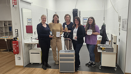 Fünf Frauen stehen zusammen in einem Raum der für die Pflege ausgestattet ist. Sie zeigen Geräte, die das Leben im Alter technisch unterstützen können.
