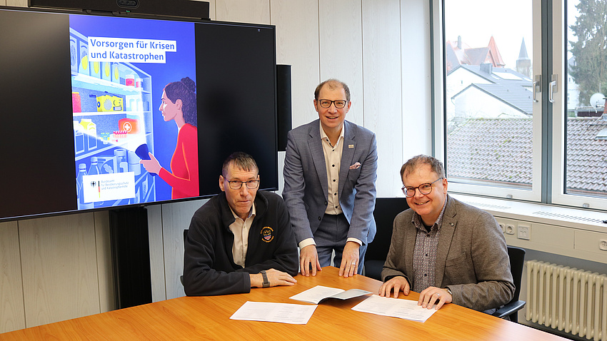 Drei Männer sitzen oder stehen an einem Tisch in einem Konferenzraum und weisen auf mehrere Broschüren vor ihnen auf dem Tisch hin. Im Hintergrund ist auf einem Bildschirm eine Grafik zu sehen.