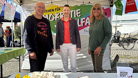 Zwei Frauen und ein Mann stehen gemeinsam an einem Infostand.