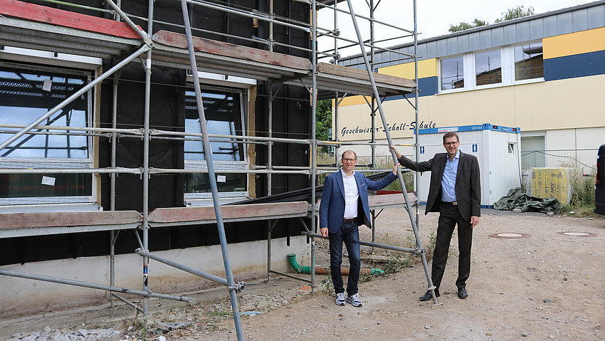Zwei Männer stehen auf einer Baustelle. Zu sehen ist ein Gebäude mit einem Baugerüst.