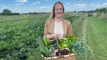 Eine Frau steht in einem Feld. Sie hält einen Gemüsekorb in die Kamera und lächelt.