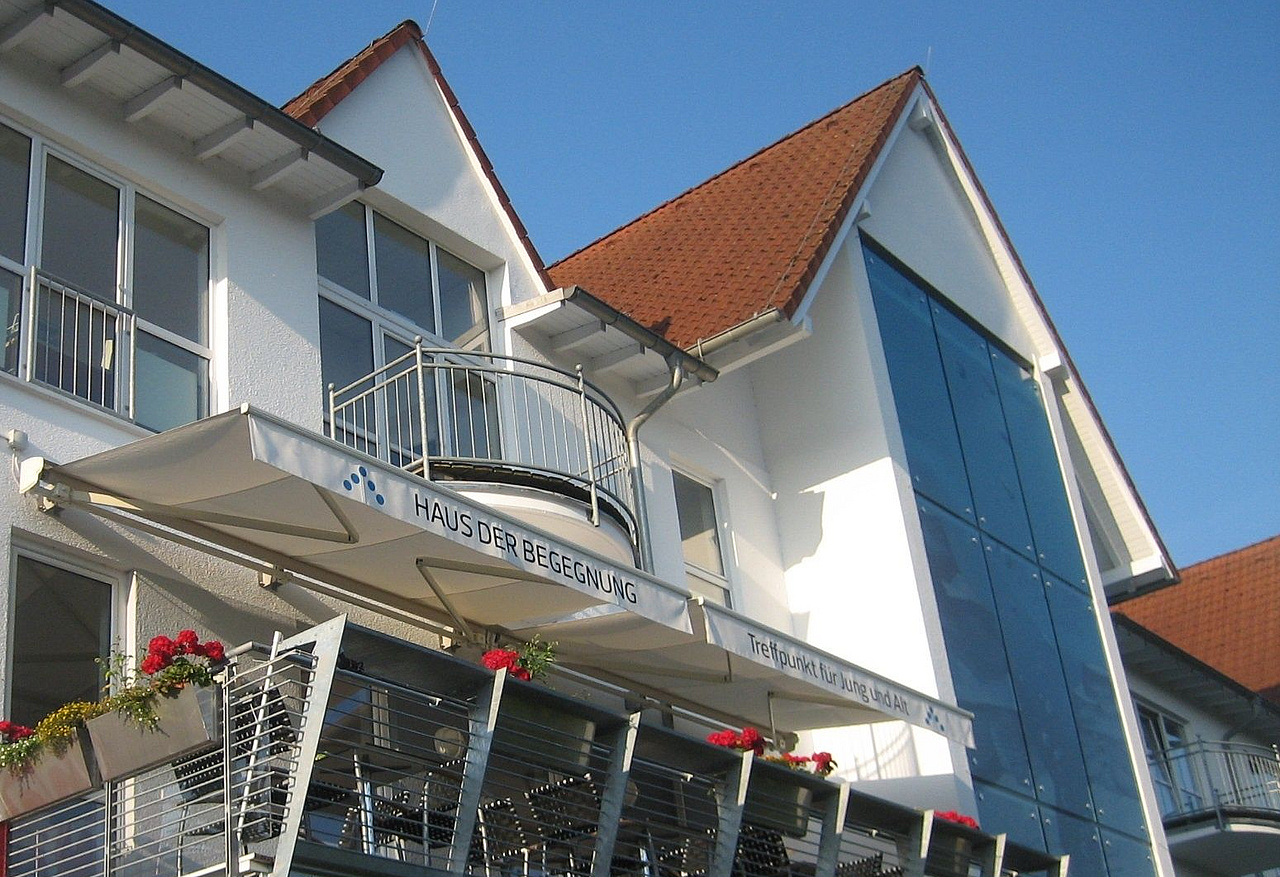 Haus mit großem Balkon, darauf Tische und Stühle sowie einer Markise mit Aufschrift „Haus der Begegnung. Treffpunkt für Jung und Alt“.