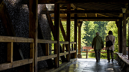 Zwei ältere Frauen gehen an der Reisig-Wand eines Gradierwerks entlang.