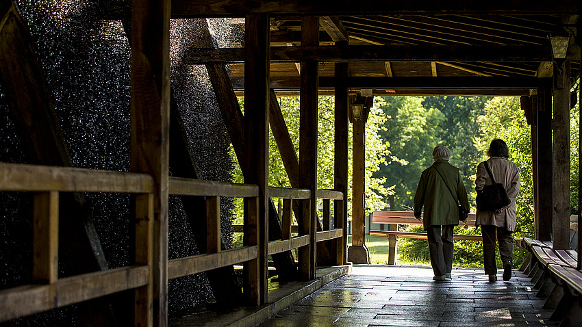 Zwei ältere Frauen gehen an der Reisig-Wand eines Gradierwerks entlang.