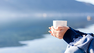Vor dem Hintergrund eines Sees wird eine weiße Tasse gehalten