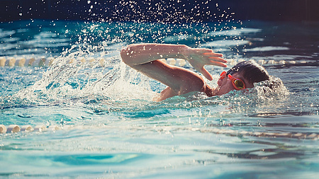 Ein Junge schwimmt im Wasser. Er krault undträgt eine Schwimmbrille.