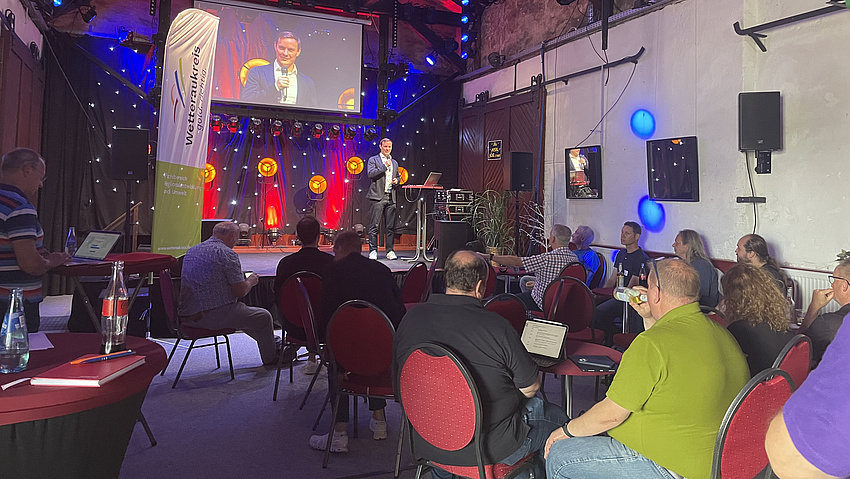 Auf der Bühne spricht ein Mensch, davor sitzen einige auf Stühlen.