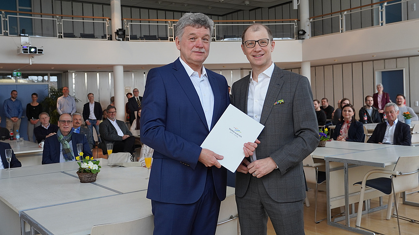 Zwei Männer stehen in einem großen Saal. Im Hintergrund sind Tische zu sehen, an denen Personen sitzen oder auch stehen. Die beiden Männer im Vordergrund halten gemeinsam ein Dokument in Händen.