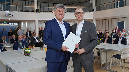 Zwei Männer stehen in einem großen Saal. Im Hintergrund sind Tische zu sehen, an denen Personen sitzen oder auch stehen. Die beiden Männer im Vordergrund halten gemeinsam ein Dokument in Händen.