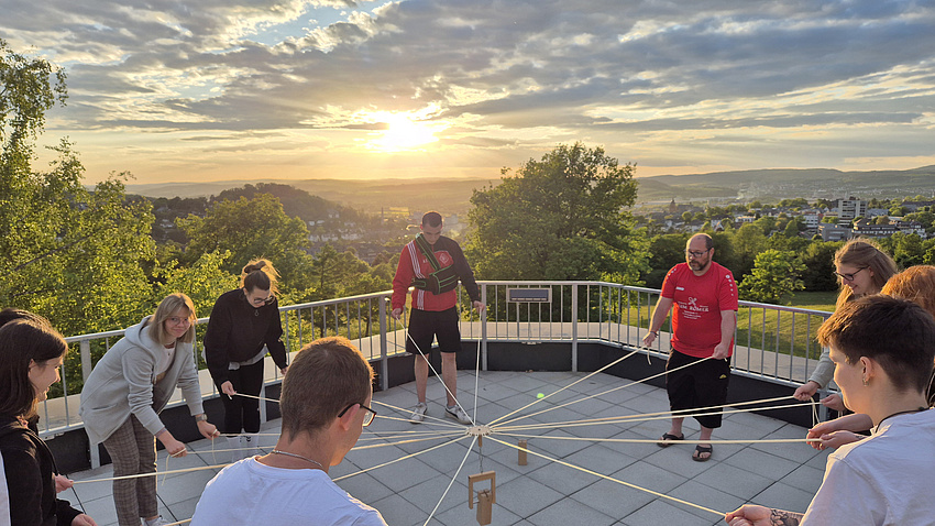 Eine Gruppe Menschen steht im Kreis aus einer Terrasse und baut gemeinschaftlich mit Seilen einen Turm.