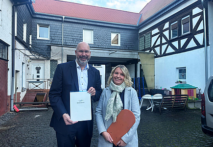 Ein Mann und eine Frau vor einem Fachwerkhaus. Der Mann hält ein Schriftstück in der Hand, die Frau einen Dachziegel.