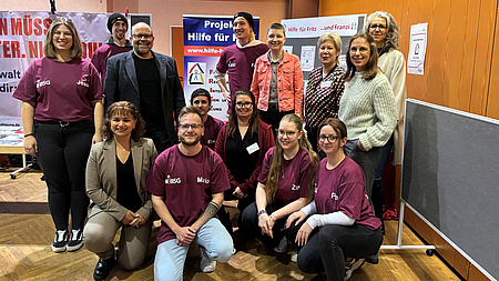 Mehrere Frauen und Männer verschiedenen Alters stehen oder knieen in der Ecke eines Saal. Im Hintergrund sind Stellwände und verschiedene Plakate zu sehen.