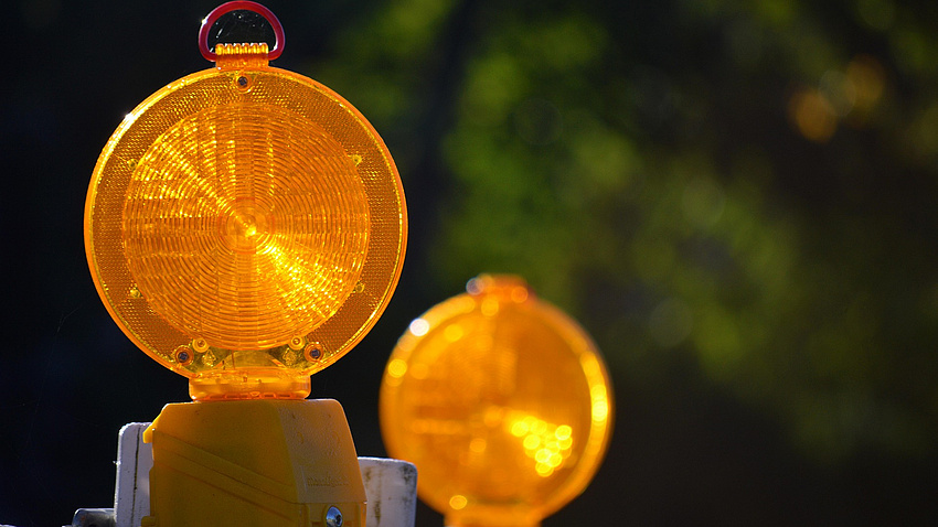 Zwei gelbe Verkehrswarnleuchten stehen in einem gewissen Abstand zueinander vor einem Hintergrund aus Pflanzen.