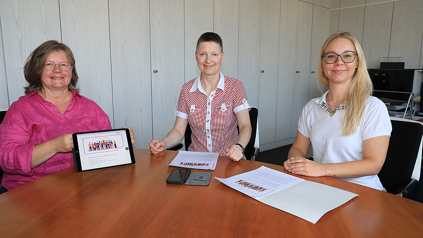 Drei Frauen sitzen in einem Büro an einem großen Konferenztisch. Eine der Frauen hält ein Tablet, die anderen beiden haben Dokumente vor sich liegen.