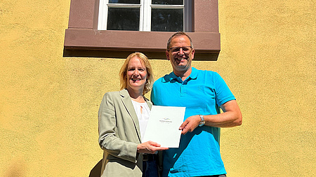 Zwei Personen stehen vor dem denkmalgeschützten Haus zur Übergabe des Förderbscheids. Das Haus hat eine gelbe Fassade und rot umrandete Fenster.