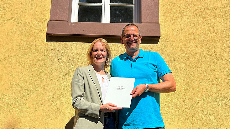 Zwei Personen stehen vor dem denkmalgeschützten Haus zur Übergabe des Förderbscheids. Das Haus hat eine gelbe Fassade und rot umrandete Fenster.