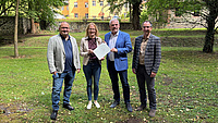 Ein Frau und drei Männer stehen in einem Park, im Hintergrund ist ein Schloss zu sehen. Die Frau und einer der Männer halten zusammen ein Dokument in Händen.